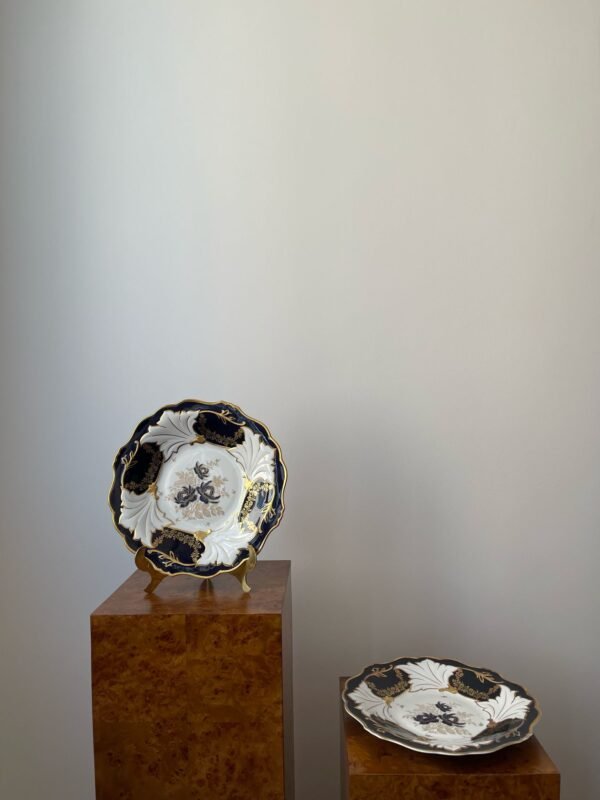 Two Weimar Echt Kobalt plates with a rich cobalt blue border and ornate gold decoration are shown, one slightly elevated on a wooden stand. The plates feature a central white area with gold and blue floral or foliate motifs.