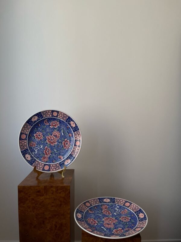 Two oval Japanese Imari porcelain plates are displayed, one upright on a stand atop a wooden pedestal, and the other lying flat on a lower pedestal. Both plates feature a vibrant blue background with detailed red floral patterns and gold accents, surrounded by intricate borders.