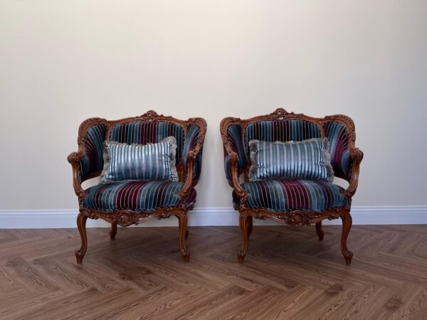 Two antique Victorian parlor chairs upholstered in striped blue, burgundy, and teal velvet, each with a matching fringed pillow, are positioned on a rug.