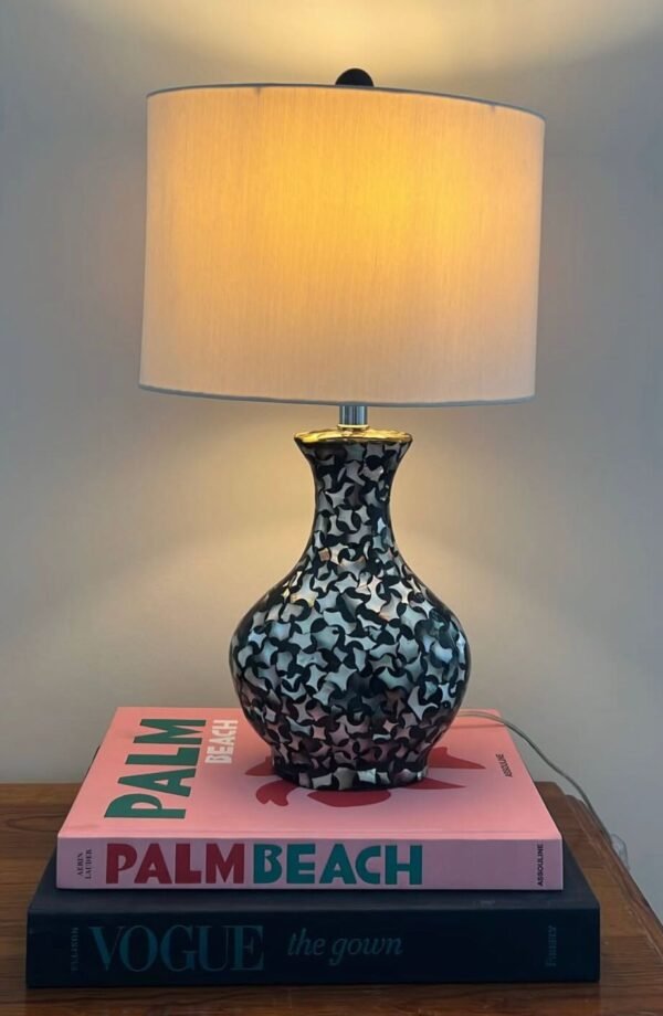 A Bloomingville table lamp with a round capiz shell base in a black, white, and grey floral or swirling pattern sits on a wooden surface, illuminated by its off-white linen drum shade.