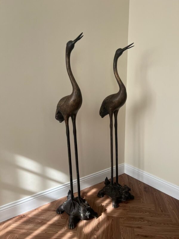 Two elegant bronze sculptures of cranes stand on a wooden floor against a light beige wall. The cranes have long legs and necks, with their heads tilted upwards. They stand on detailed, textured bases.